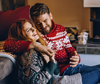 Ein mann und eine Frau sitzen auf dem Boden vor Ihrem Sofa mit weihnachtlichen Pullovers. Die Frau lacht, während der Mann Ihr gerade etwas erzählt.