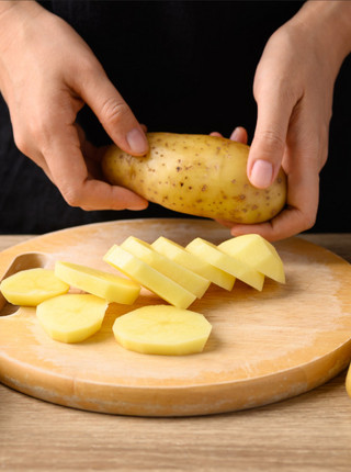 Kartoffelscheiben liegen auf einem Holzbrett, während eine Hand eine Kartoffel hält.