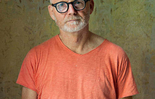 Ein Mann mit weißem Bart, schwarzer Brille und einer Glatz steht vor einer gemusterten Tapete für ein Portraitfoto