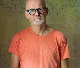 Ein Mann mit weißem Bart, schwarzer Brille und einer Glatz steht vor einer gemusterten Tapete für ein Portraitfoto