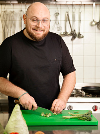 Michael Hergenröder schneidet gerade Gemüse, während er lächelnd in die Kamera schaut.