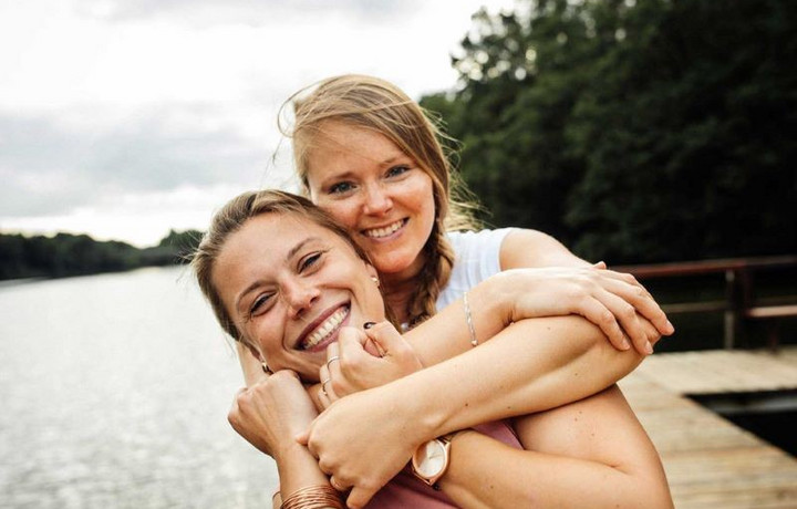 Zwei Frauen umarmen sich an einem See und blicken in die Kamera