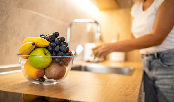 Eine Frau wäscht sich im Hintergrund die Hände an der Küchenspüle, während sich im Vordergrund ein Glasschüssel voller Obst befindet.