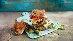 Ein Gourmet-Sandwich mit Speck, geschmolzenem Käse und Garnitur auf einem rustikalen Holzbrett, dazu Gurken.