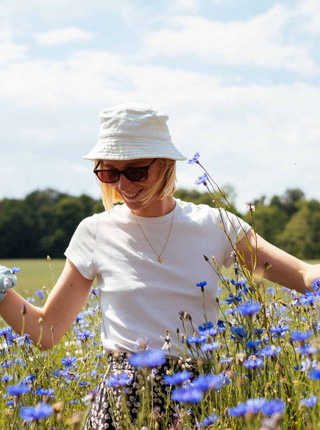 Junge Frau mit weißem T-Shirt, weißem Hut und Sonnenbrille in einem Meer aus blauen Kornblumen