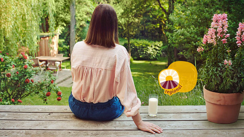 Das Symbolbild steht für regionale Produkte und zeigt eine Frau sitzend auf einer Holzterrasse mit einem Glas Milch in der Hand.