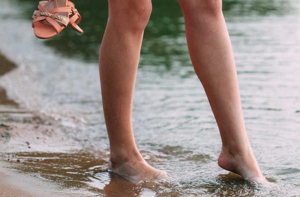 Frau mit Füßen im Wasser und Schuhen in der Hand