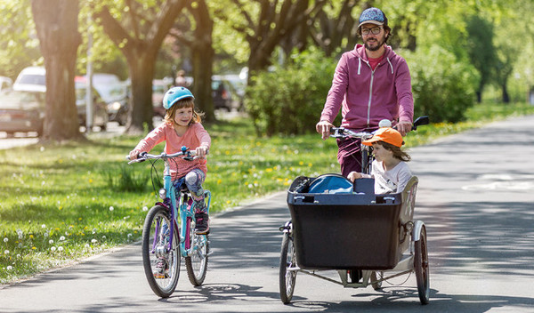 Ein Vater fährt mit seinen zwei Kindern Fahrrad. Das Eine Kind sitzt im Lastenfahrrad.