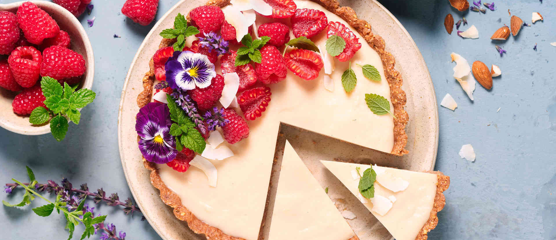veganer Kuchen als Tarte mit Himbeeren und lila Blüten dekoriert vor einem hellblauen Hintergrund