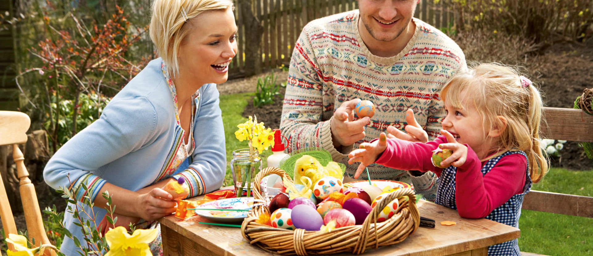 Eine Mutter, ein Vater und Ihr Kind sitzen zusammen an einem Holztisch im Garten und bemalen gemeinsam Ostereier