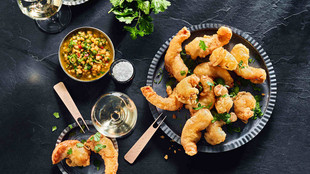 Teller mit knusprig frittierten Garnelen mit Dip-Sauce und Gläsern Weißwein auf einer dunklen Tischplatte, garniert mit frischen Kräutern und Meersalzflocken.