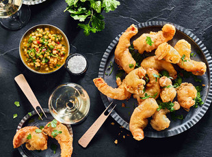 Teller mit knusprig frittierten Garnelen mit Dip-Sauce und Gläsern Weißwein auf einer dunklen Tischplatte, garniert mit frischen Kräutern und Meersalzflocken.