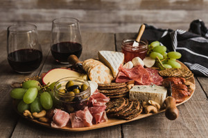 Unterschiedliche Lebensmittel wie Salami, Schinken, Käse, Oliven auf Brettchen angerichtet, im Hintergrund Weingläser.