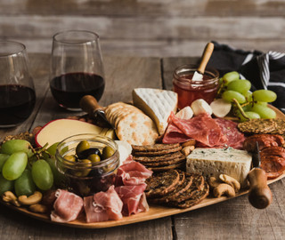 Unterschiedliche Lebensmittel wie Salami, Schinken, Käse, Oliven auf Brettchen angerichtet, im Hintergrund Weingläser.