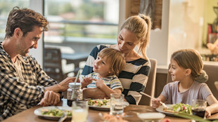 Eine Familie sitz am Essenstisch und schauen, während sie essen, ihr jüngstes Kind an, was auf dem Schoß der Mutter sitzt.