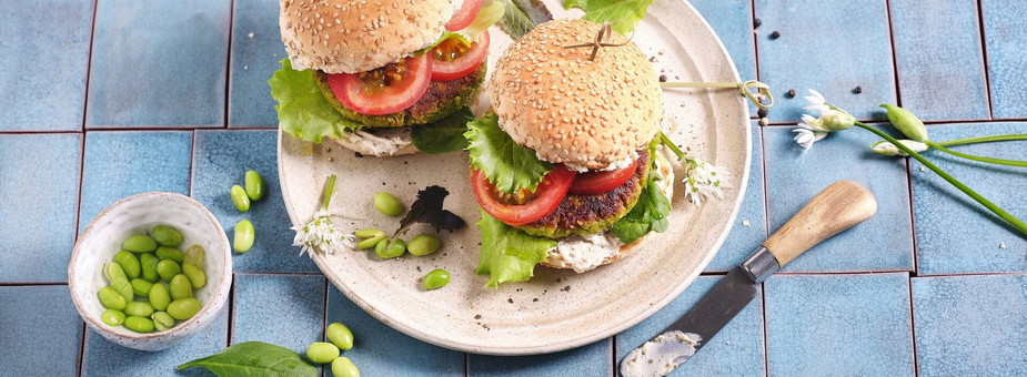 Zwei leckere Burger mit veganem Protein Patty, Tomaten und Salat an grünen Edamame Bohnen auf einem weißen Teller.