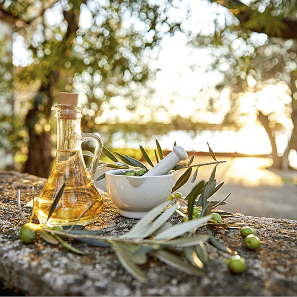 Ein Olivenflasche, ein Mörser und Olivenbaumzweigen stehen auf einem steinigen Untergrund, während im Hintergrund die Sonne untergeht