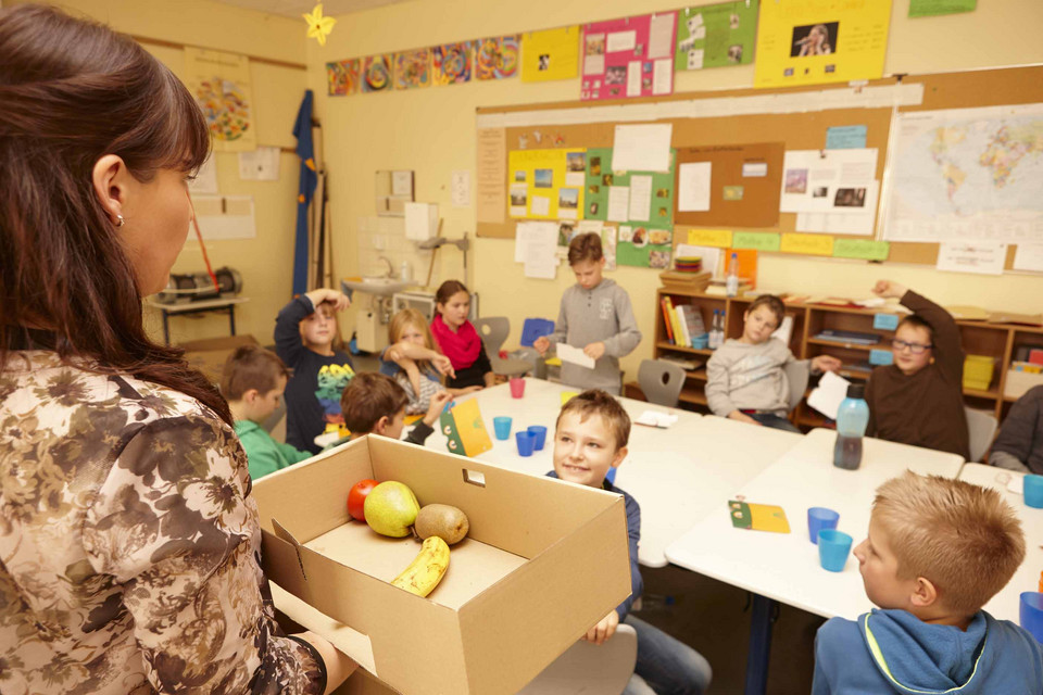 Frau steht mit Karton mit Obst in einem Klassenraum mit Schülern