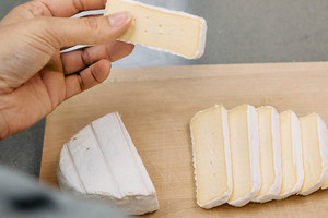 Ein Stück Camembert wird in der Hand gehalten, während der Rest auf einem Brett angerichtet wurde.