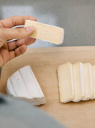 Ein Stück Camembert wird in der Hand gehalten, während der Rest auf einem Brett angerichtet wurde.