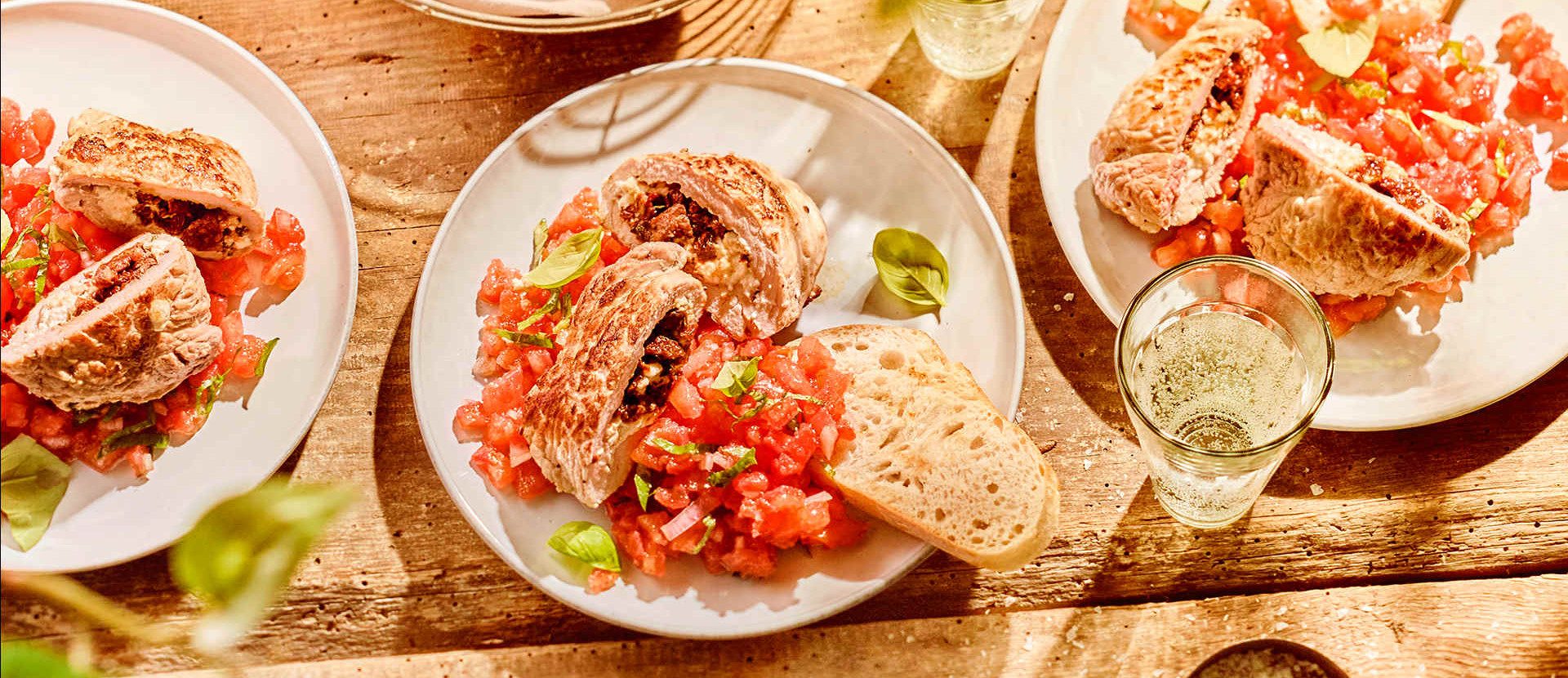 Draufsicht auf einen rustikalen Holztisch mit Tellern voller Bruschetta, belegt mit Tomaten und Basilikum, Brotscheiben und Gläsern Weißwein im Sonnenlicht.