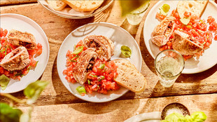 Draufsicht auf einen rustikalen Holztisch mit Tellern voller Bruschetta, belegt mit Tomaten und Basilikum, Brotscheiben und Gläsern Weißwein im Sonnenlicht.