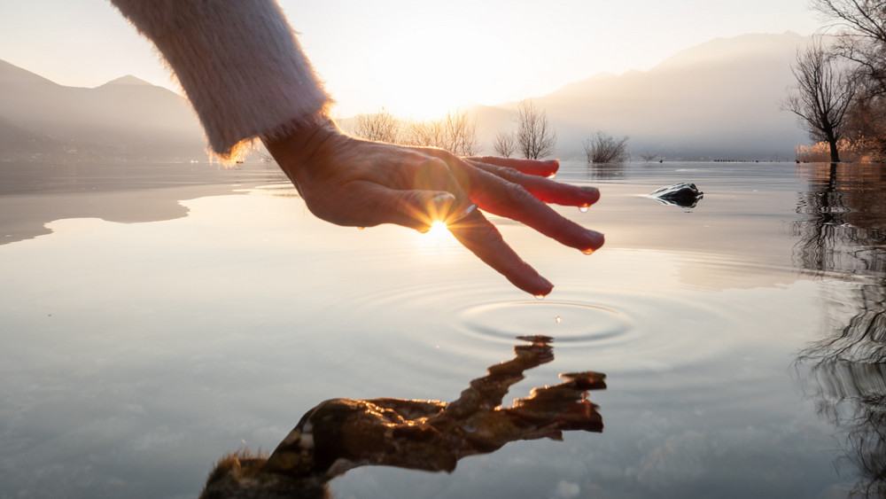 Eine Hand berührt die Wasseroberfläche des Sees bei Sonnenuntergang. Die Hand spiegelt sich in der Wasseroberfläche.
