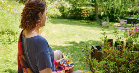 Eine Frau sitzt im Schneidersitz im Garten, während sie eine Schüssel mit Müsli in der Hand hält