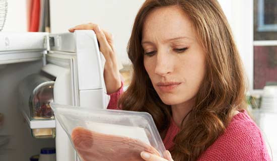Eine dunkelhaarige Frau kontrolliert das Mindesthaltbarkeitsdatum einer Schinkenpackung vor dem geöffneten Kühlschrank.