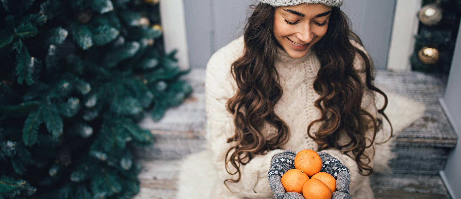 Eine junge Frau sitzt neben einem Weihnachtsbaum und hält vier Mandarinen in ihrer Hand, während sie diese anschaut