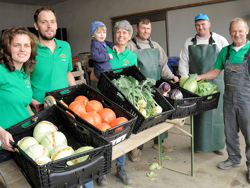 Sechs Mitarbeiter und Mitarbeiterinnen präsentieren die Gemüseernte, eine Frau hält ein Kind auf dem Arm