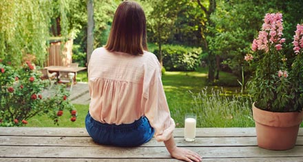 Zu sehen ist eine dunkelhaarige Frau von hinten, die auf einer Holzterrasse sitzt, daneben ein Glas Milch.