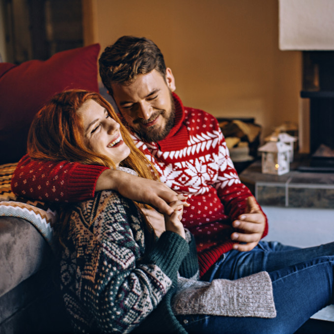 Ein mann und eine Frau sitzen auf dem Boden vor Ihrem Sofa mit weihnachtlichen Pullovers. Die Frau lacht, während der Mann Ihr gerade etwas erzählt.