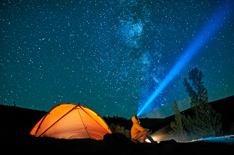 Person mit Stirnleuchte sitzt in sternklarer Nacht vor einem Zelt und blickt in den Himmel.